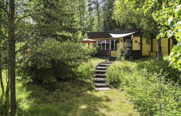 Ein gelbes Holzhaus umgeben von Bäumen, mit einer Treppe und grünem Gras davor.